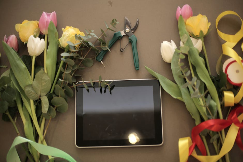 A flat lay of fresh flower bouquets featuring pink, yellow, and white tulips with green leaves and stems, arranged on a neutral surface. The flowers are positioned on either side of a black tablet, wh