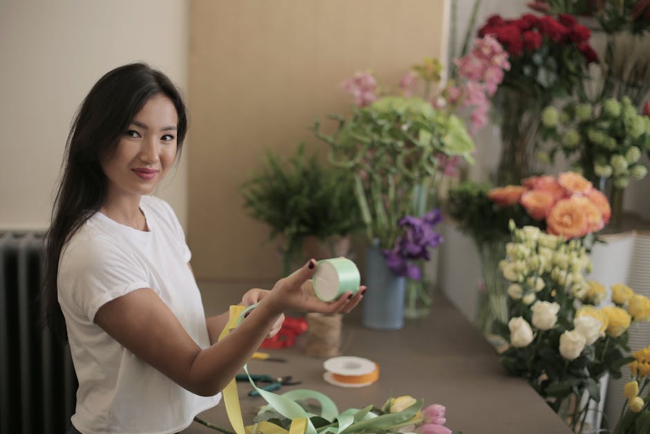 A young woman with long dark hair and a white shirt is smiling while holding a roll of light green ribbon in her right hand and a pair of yellow scissors in her left hand. She is standing in a floral 