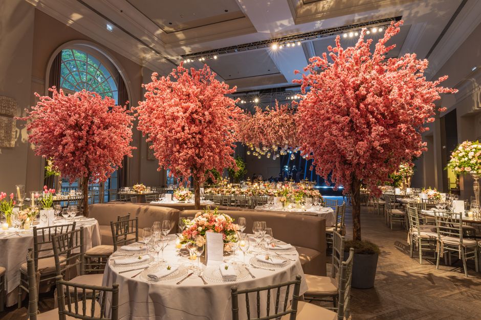 An elegant banquet hall decorated with large, vibrant pink cherry blossom trees placed at the center of round tables, which are draped with white tablecloths and set with fine glassware, silverware, a