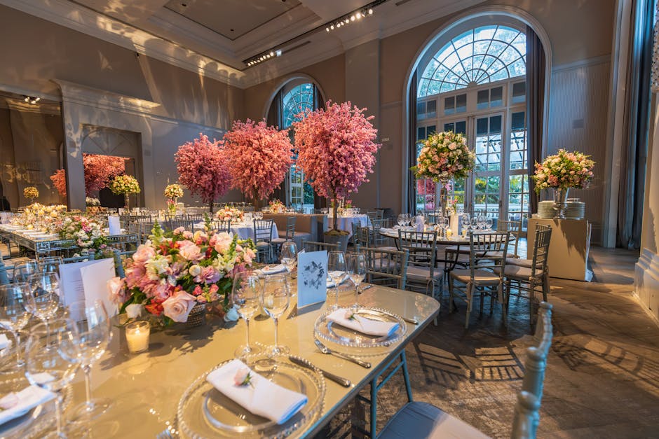 Elegant interior of a banquet hall decorated with floral arrangements, featuring large pink cherry blossom trees and smaller bouquets of pink and white roses with greenery, placed in tall glass vases 