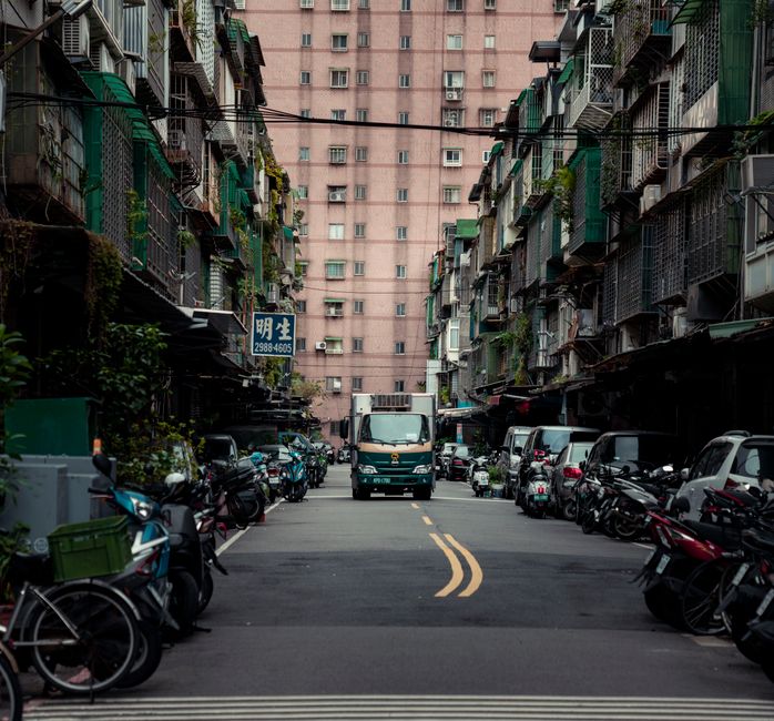 The image depicts a narrow street in an urban setting, flanked by multi-story residential buildings with balconies, some of which have green awnings and potted plants. Parked motorcycles and bicycles