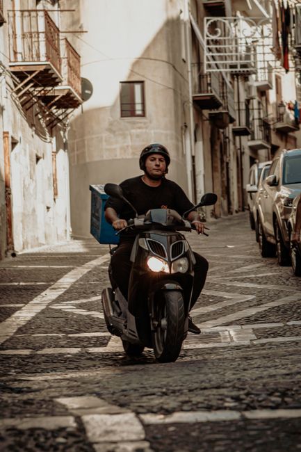 A delivery person riding a black scooter through a narrow, cobbled street lined with old multi-story buildings featuring external fire escapes and balconies, carrying a package on their back. The scen