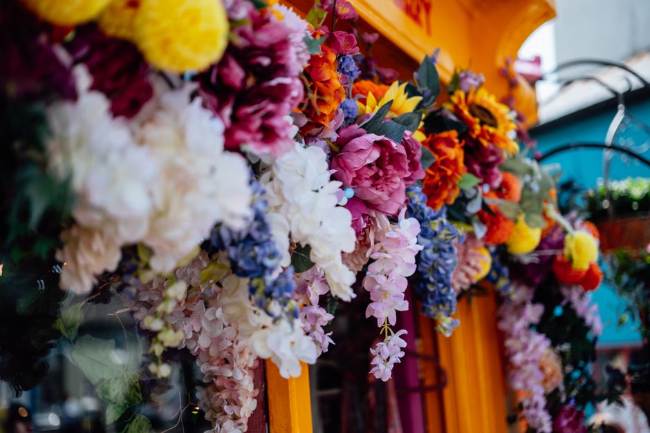 A vibrant floral display featuring a variety of fresh flowers such as pink peonies, white hydrangeas, yellow sunflowers, orange marigolds, purple delphiniums, and red roses. The arrangement is densely