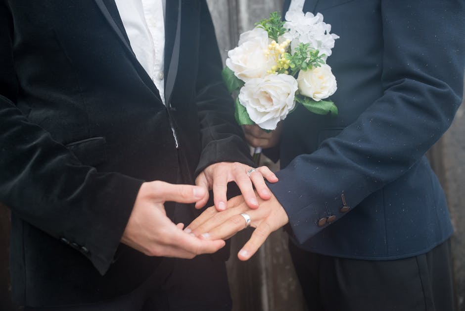 A close-up of a person in a dark suit holding a bouquet of fresh white flowers, including roses and hydrangeas, wrapped in clear cellophane and a green accent foliage. The flowers are arranged neatly