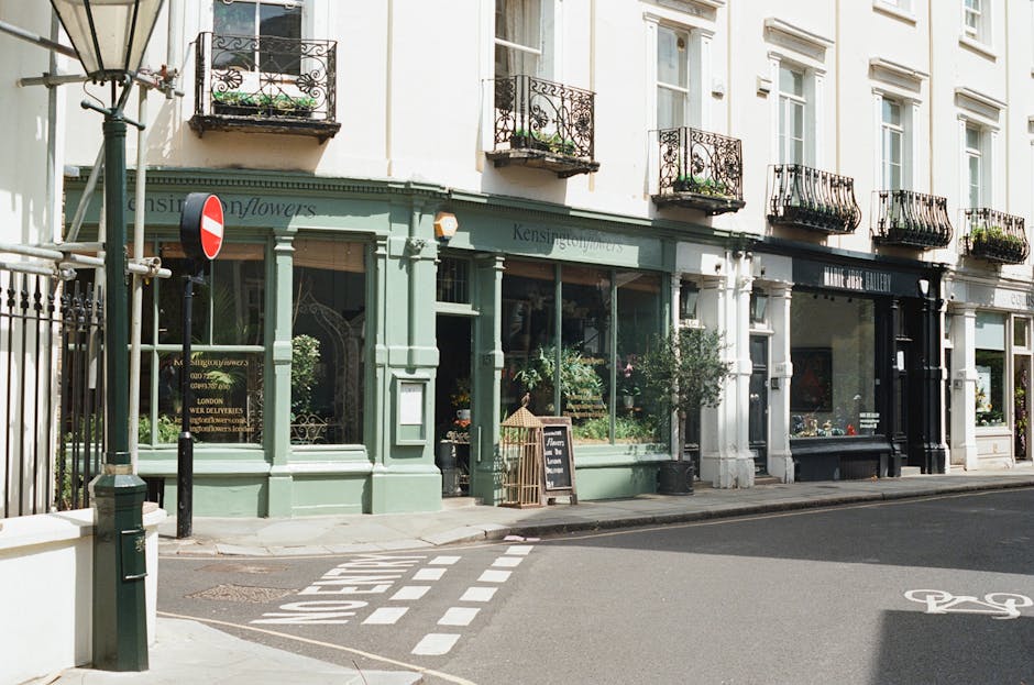 The exterior of a floral shop on a city street, featuring a light green storefront with large glass windows displaying an array of fresh flowers and potted plants inside. The shop has decorative black