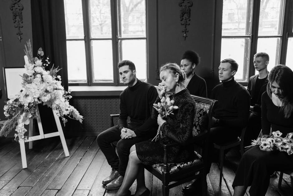 A black-and-white photograph showing a group of six individuals seated in a room with large, multi-pane windows. The central woman is holding a bouquet of tulips and appears to be reflecting or mourni