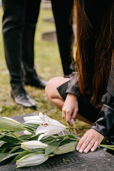 A person with long brown hair, dressed in black, is kneeling and carefully placing a white lily among a bouquet of similar lilies with green leaves, arranged on a stone surface. The lilies have large,