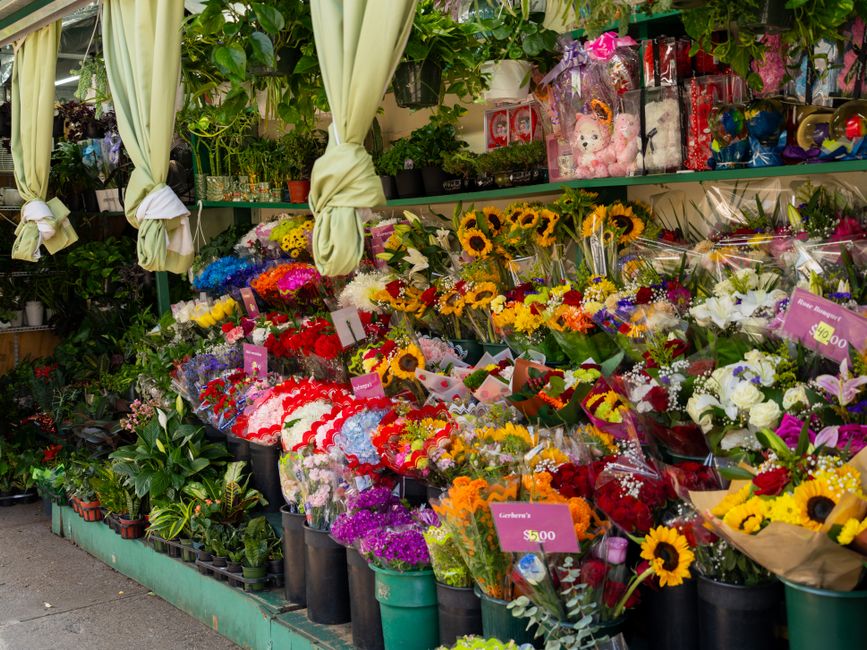 A vibrant flower stall displaying an assortment of fresh floral arrangements arranged in black buckets and wrapped bouquets. The flowers include bright yellow sunflowers, red roses, purple statice, wh