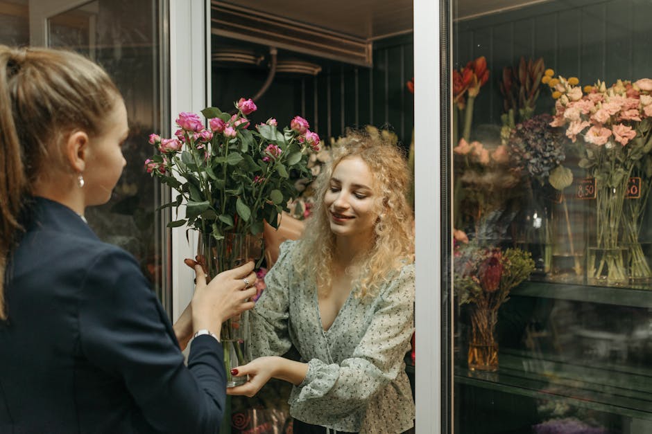 A florist at Mayfair, wearing a dark blazer, is handing a tall glass vase filled with fresh, vibrant pink roses to a smiling woman with curly blonde hair, dressed in a floral patterned blouse. The wom