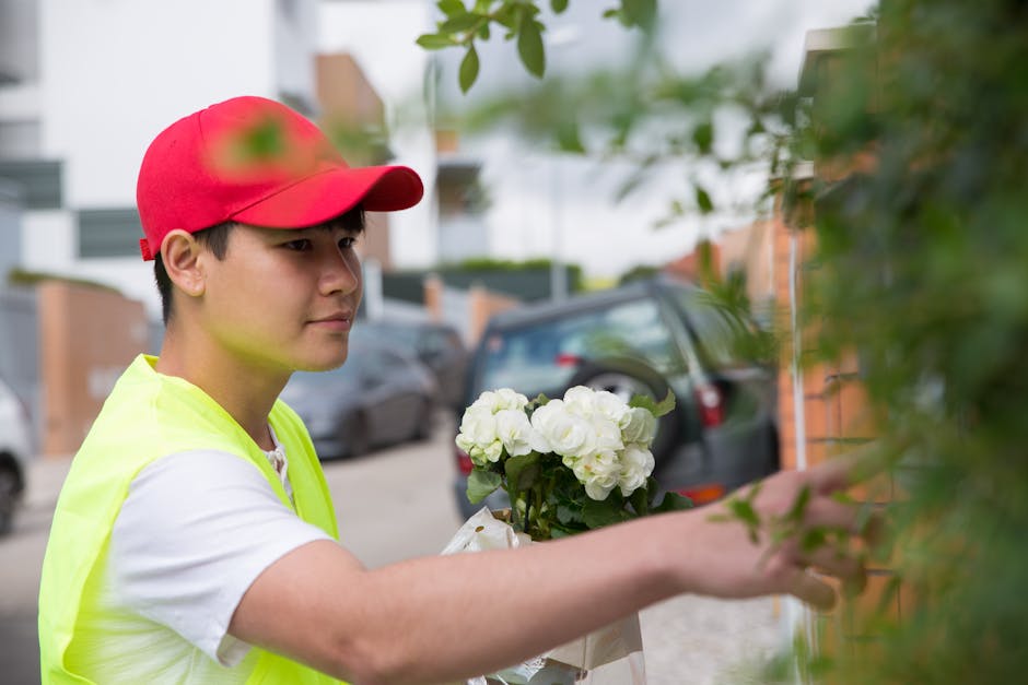 A young male florist wearing a red cap and a neon yellow vest is arranging a bouquet of fresh white flowers, possibly hydrangeas or roses, wrapped in white paper. He is standing outdoors near a reside